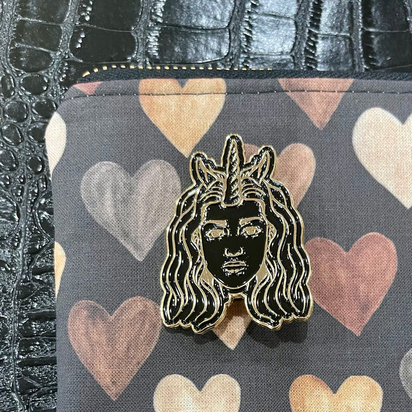 Half Unicorn enamel pin of the company's logo in black and gold. Features woman with wavy hair and a unicorn horn as well as horse-like ears. Pin on corner of a pouch with multi-colored heart fabric design.