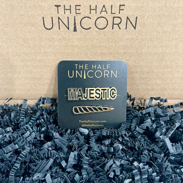 Two enamel pins: the word Majestic  and a unicorn horn, both in black and gold and attached to a small black card with The Half Unicorn's business logo in gold on it. The set  sits on black crinkle paper in a brown Kraft box stamped with the company logo.