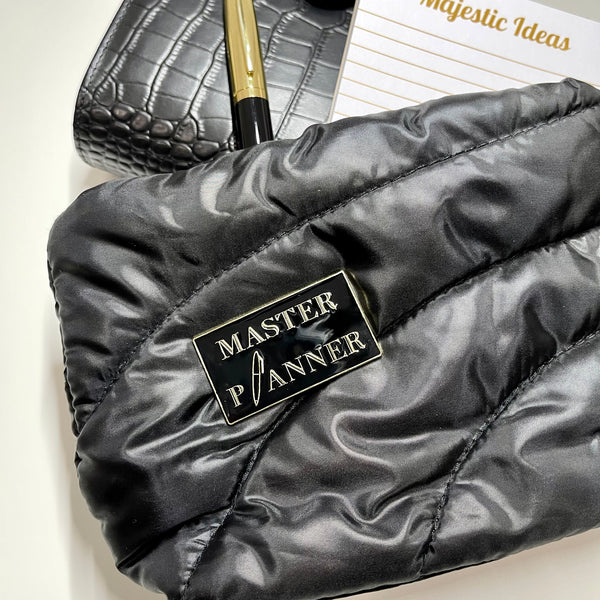 Master Planner enamel pin in black and gold colors attached to a black quilted pouch. A black croc planner, gold pen, and notepad are sticking out of the pouch.