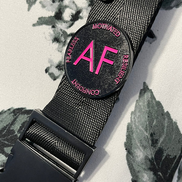 As F**k enamel pin in pink and black colors attached to a black backpack strap.