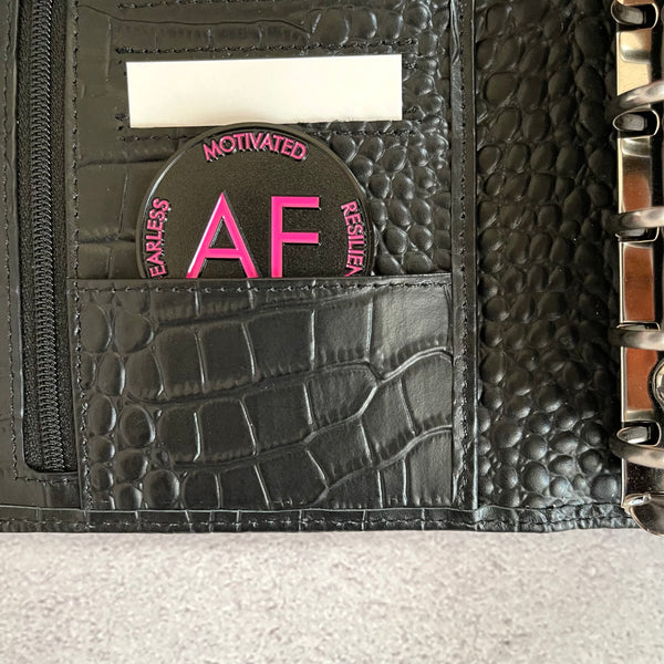 As F**k Token in pink and black colors sits inside the inner credit card slot pocket of a black croc planner.