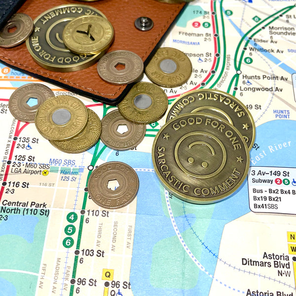 Good For One Sarcastic Comment tokens in a burnished gold color designed to look like an old subway token and some old NYC tokens scattered on top of a subway map.