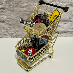 A gold miniature shopping cart filled with several different enamel pins from The Half Unicorn's shop.