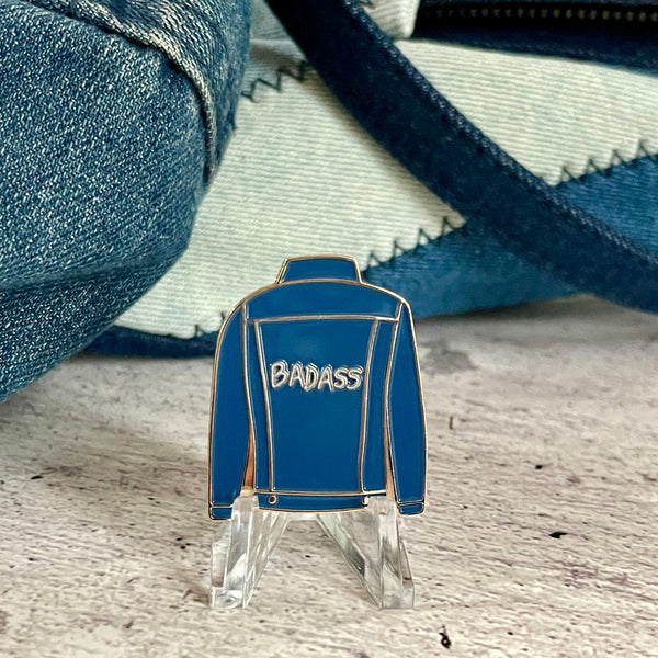 Badass Denim Jacket enamel pin sitting on a mini easel. Pin looks like the back of a blue denim jacket with the word Badass written on it.