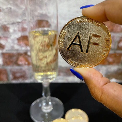 Champagne colored As F**k Token held in front of a glass of champagne.