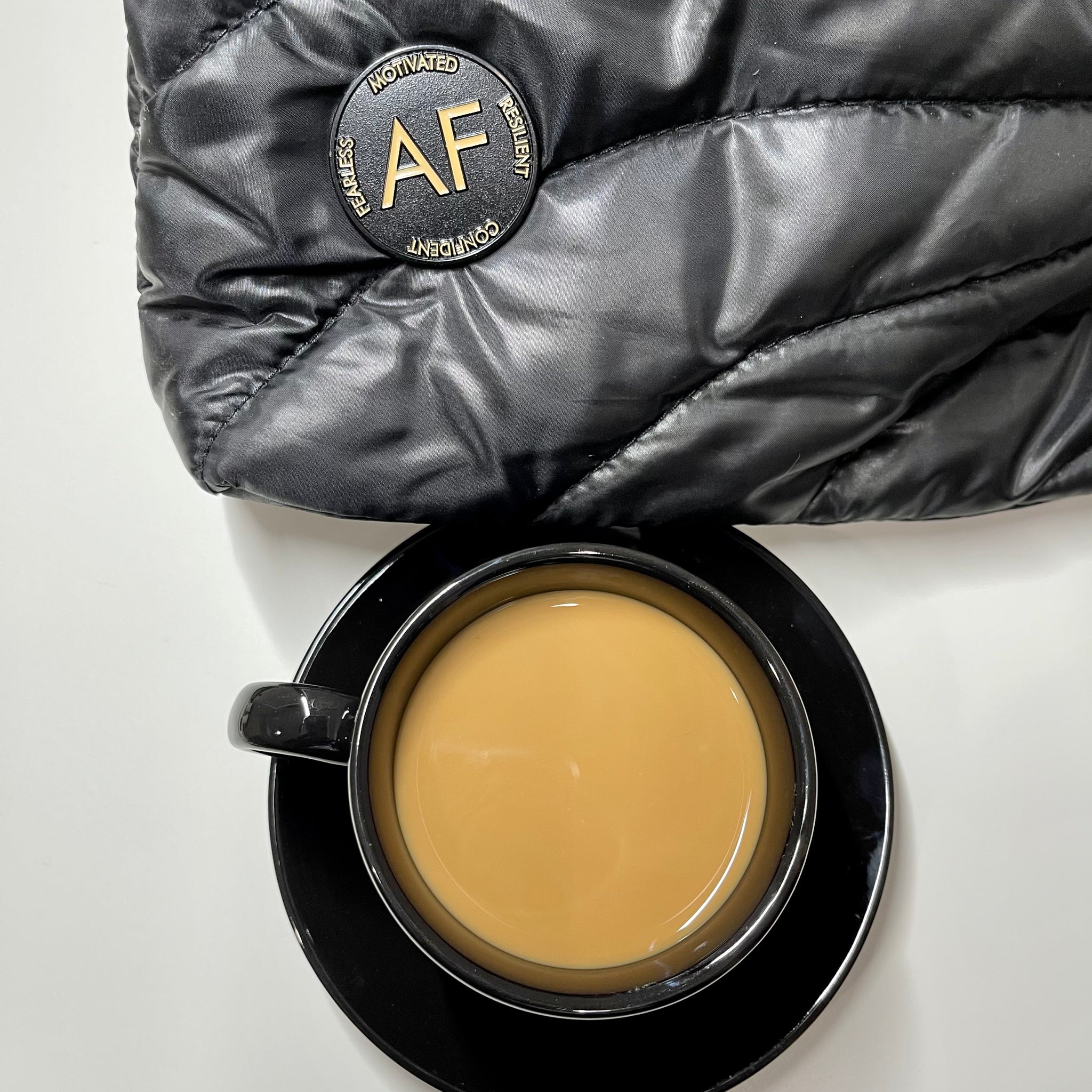 Coffee and Cream colored As F**k Enamel Pin on a black quilted pouch that is sitting by a cup of coffee.