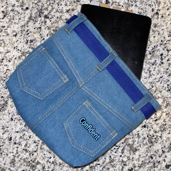 Confident enamel word pin on denim pouch with planner sticking out. Pin is cornflower blue color.