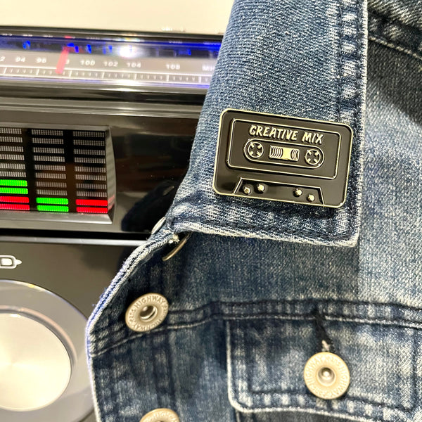 Black and gold colored Creative Mix cassette tape enamel pin on collar of a denim jacket. 