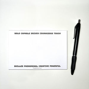 Notepad with uplifting words like BOLD, CAPABLE, DRIVEN, SKILLED, POWERFUL at the top and bottom borders. White paper with black words. Black prop pen beside notepad.