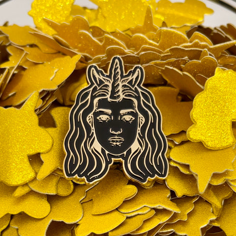 Half Unicorn enamel pin of the company's logo in black and gold. Features woman with wavy hair and a unicorn horn as well as horse-like ears. Pin sitting atop pile of gold unicorn-shaped confetti.