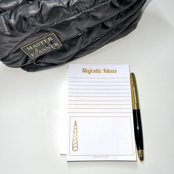 Notepad with Majestic Ideas at top, lines for writing, then a box with a unicorn horn image at the bottom. Paper is white and all lettering and images are in gold. Black and gold pen and black pouch with Master Planner enamel pin attached are props sitting by notepad.