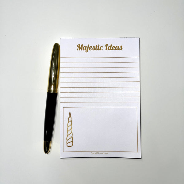 Notepad with Majestic Ideas at top, lines for writing, then a box with a unicorn horn image at the bottom. Paper is white and all lettering and images are in gold. Black and gold pen prop sits beside notepad.