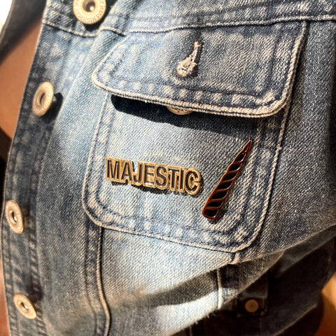 Two enamel pins: the word Majestic and a unicorn horn, both in black and gold, are attached to the pocket of a denim jacket.