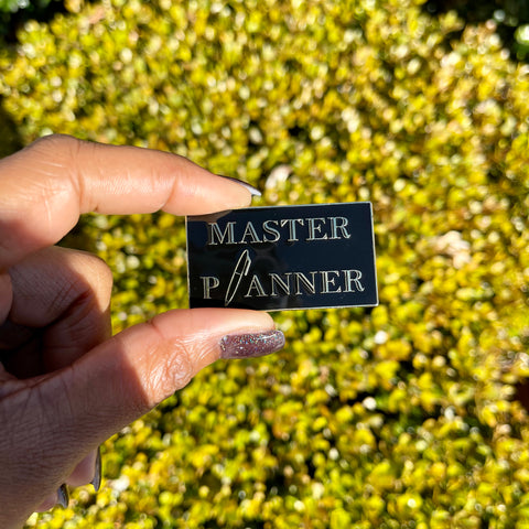 Master Planner enamel pin in black and gold colors. Pin is being held outside in front of bushes.