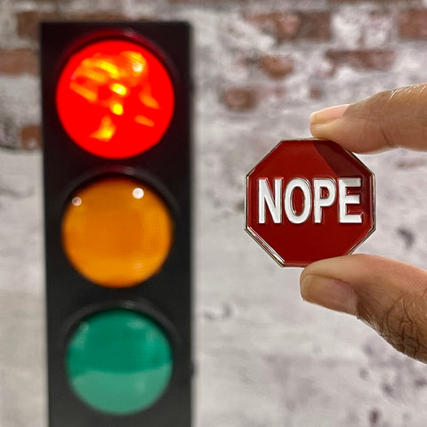 Nope Sign enamel pin that looks like a Stop Sign. It's being held beside a traffic light. The red light is lit up.