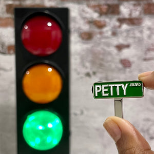 Petty Boulevard enamel pin that looks like a traditional green street sign. Pin is being held in front of a traffic light with the green light lit up.