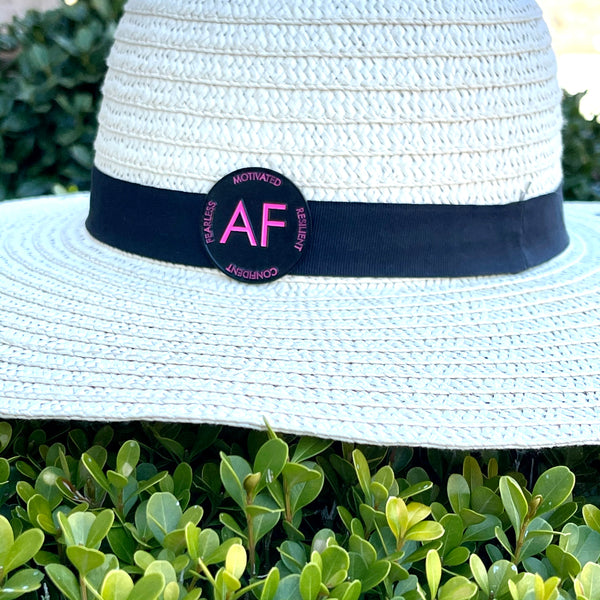 As F**k enamel pin in pink and black colors attached to a straw sun hat.  The hat sits atop a bush.