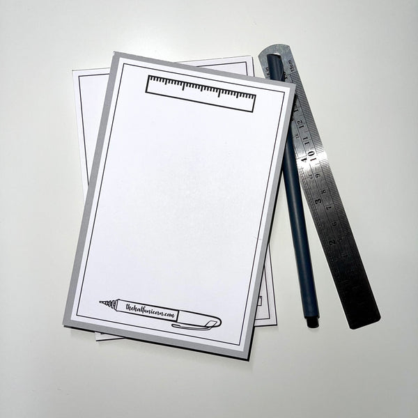Notepad with ruler image up top, pen image at bottom (pen has The Half Unicorn on the barrel). White paper with light gray border. Gray pen and ruler as props sit beside notepad.