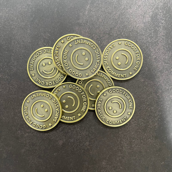 Pile of Good For One Sarcastic Comment tokens in a burnished gold color designed to look like an old subway token. Upside down smile in center of token.