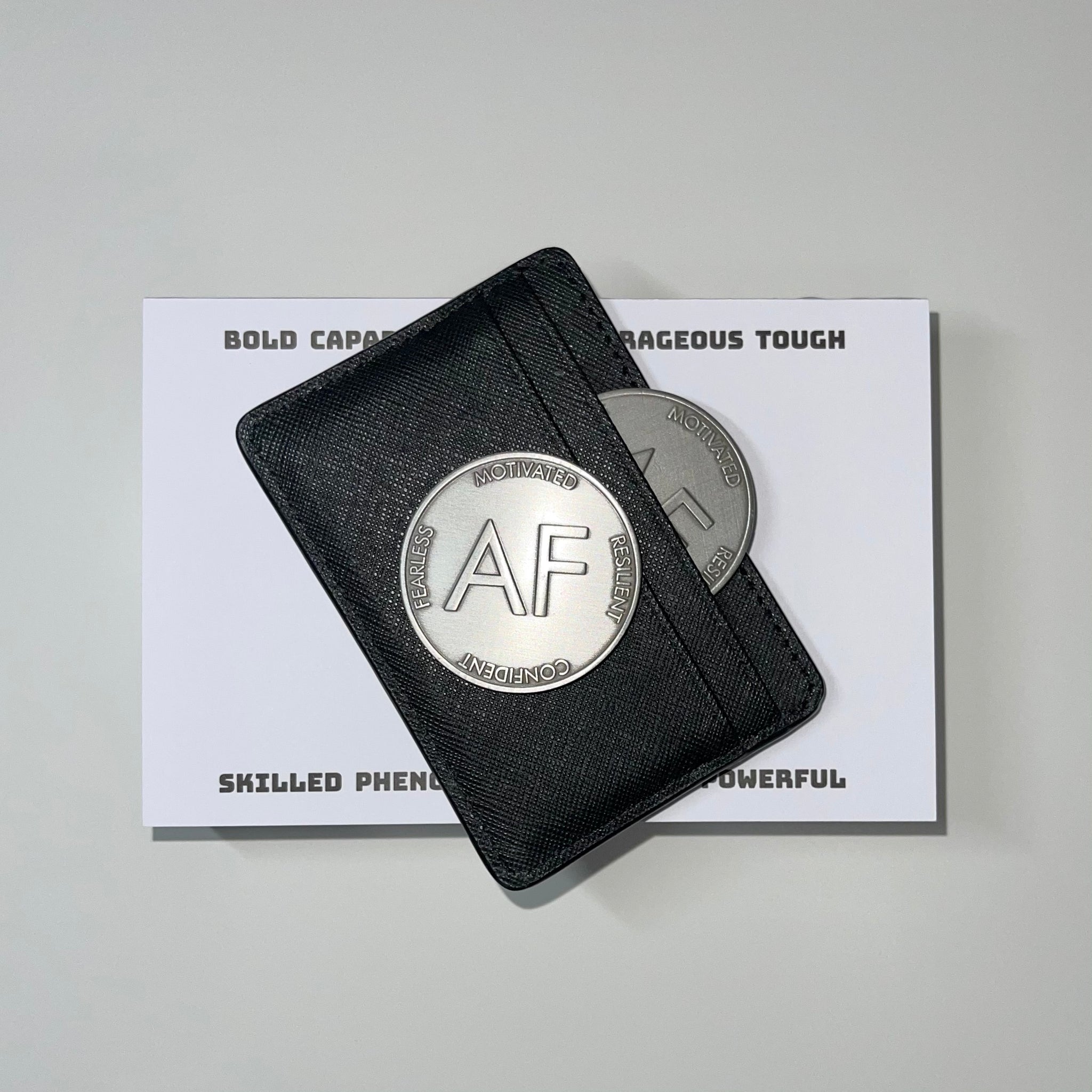 As F**K Token in worn silver color sits on a black card case while a second silver token sits partway inside the card case. The case sits on top of a notepad.