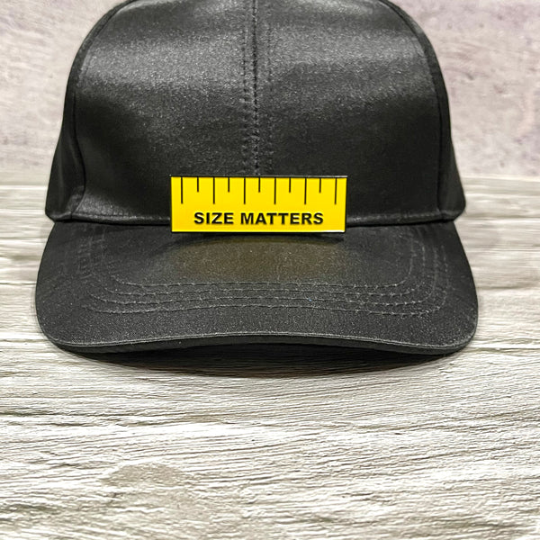 Size Matters enamel pin is yellow with black lettering and dash marks. Pin sits on the brim of a black baseball cap.