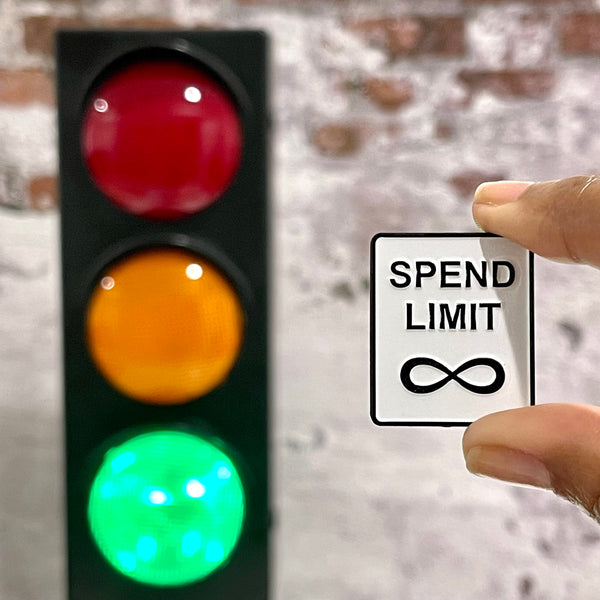 Spend Limit Sign enamel pin looks like a black and white Speed Limit Sign. Pin is being held in front of a traffic light with the green light lit up.