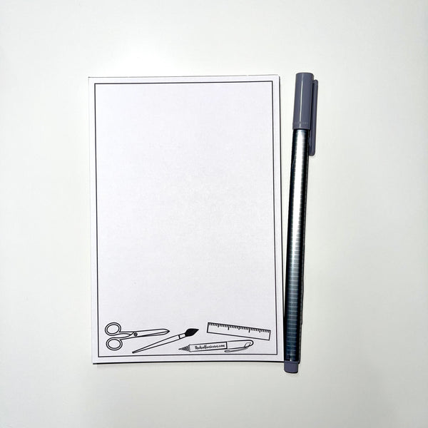 Supplies notepad has images of scissors, paint brush, ruler and a pen at the bottom. White paper with images in black.  A gray pen sits beside the notepad as a prop.