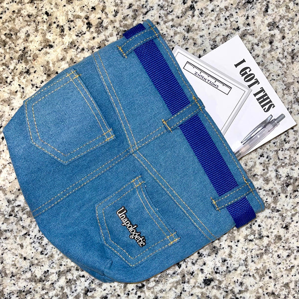 Unapologetic word enamel pin (in white) is attached to a denim pouch with two notepads poking out of the pouch as props.