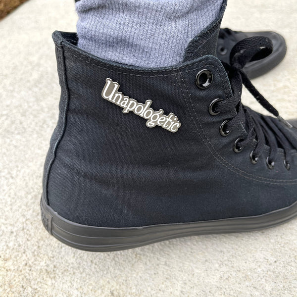 Unapologetic word enamel pin is white. Pin is attached to a black hi-top Converse sneaker.