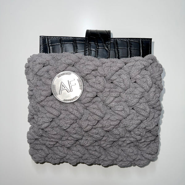 As F**k Enamel Pin in worn silver color is pinned to a gray crotchet pouch with a black planner sticking out of the pouch.